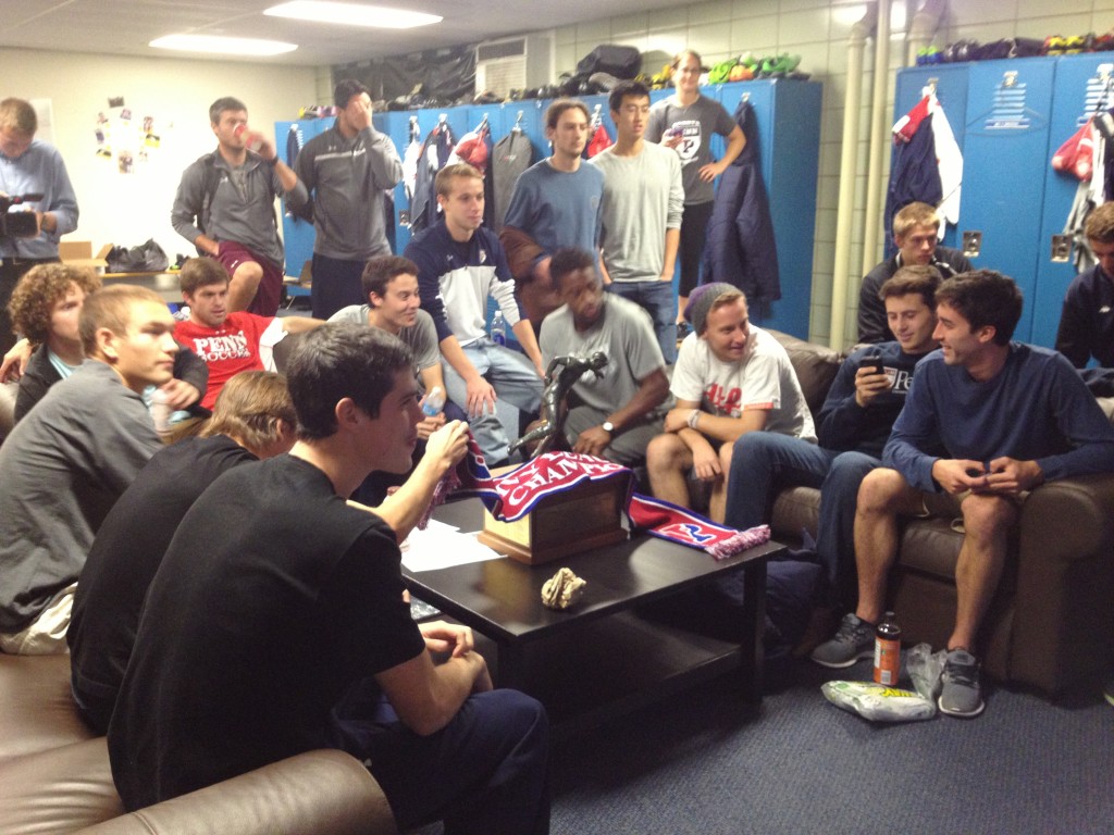 Penn soccer players and coaches watch the NCAA selection show, with their Ivy League championship trophy sitting on the table beside them.