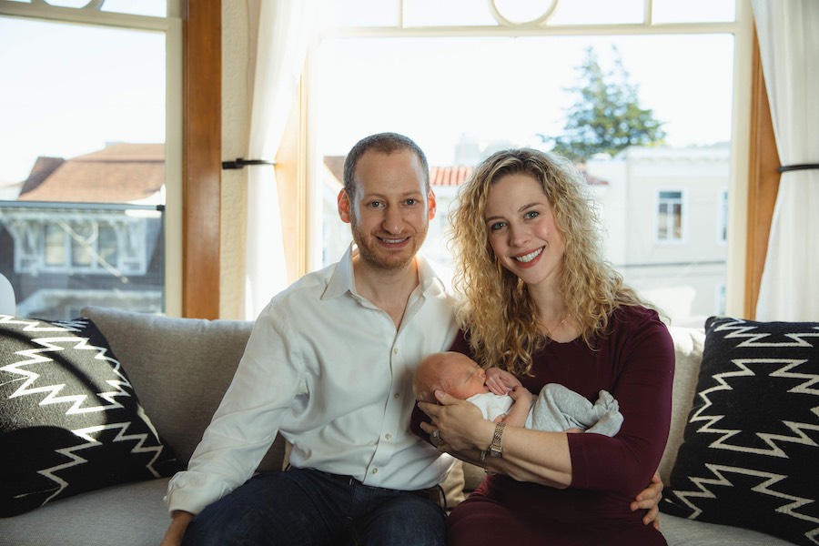 Olivier and Erica sit on a couch, holding their newborn baby.