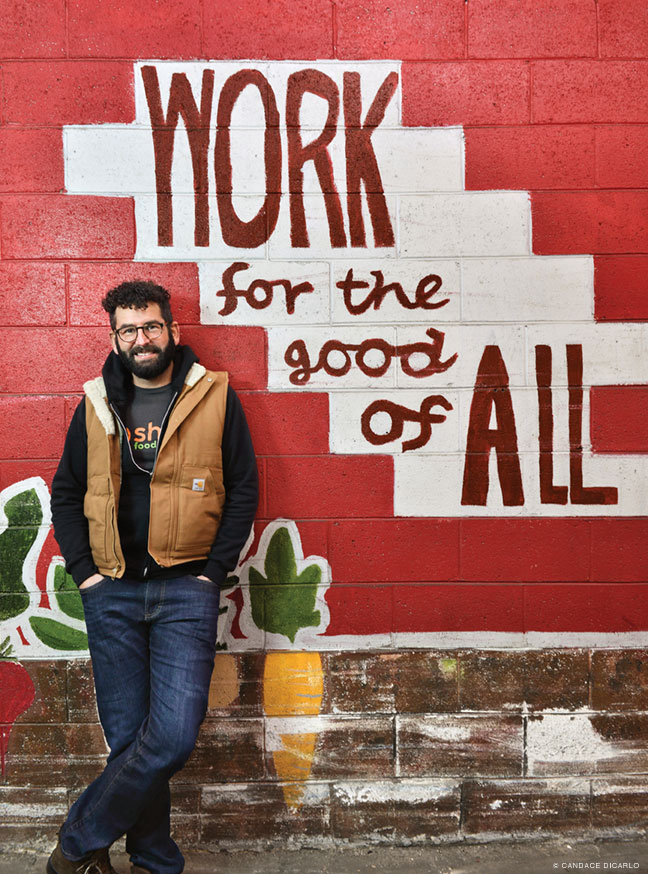 Photo of George Matysik standing in front of a mural that reads "Work for the good of All"